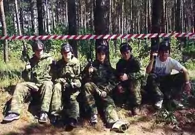 Пять человек в камуфляже и гражданской одежде сидят на земле в лесу, за ними — красно-белая предупредительная лента.