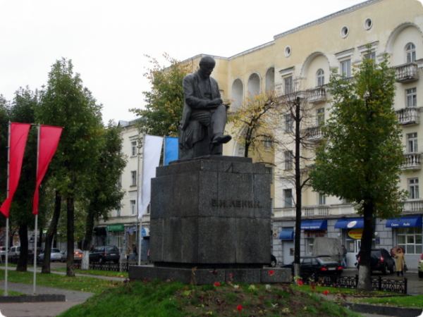 Бронзовый памятник сидящему мужчине на высоком каменном постаменте с надписью «В.И. Ленин» на фоне здания с арочными окнами и деревьев.