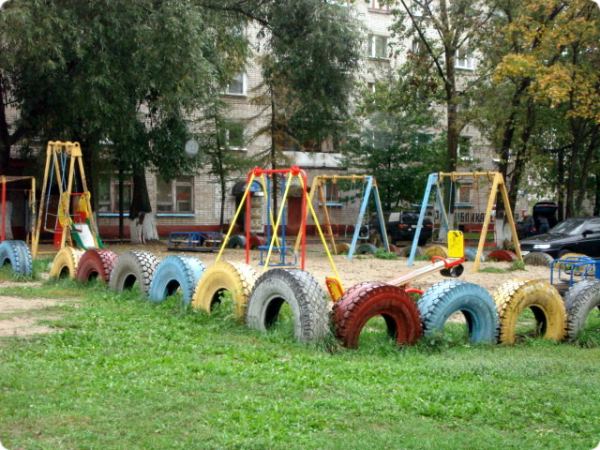 Детская площадка с качелями и качалкой-балансином, огороженная рядом разноцветных покрышек, на фоне жилого дома и деревьев.