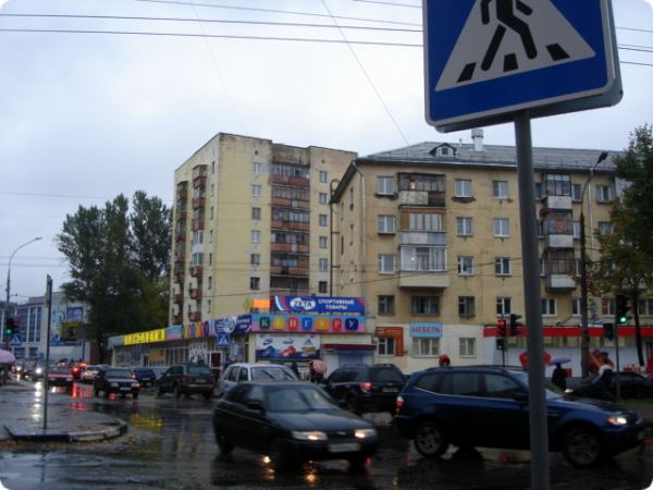 Мокрая городская улица с движущимися автомобилями, пешеходным переходом и жилыми домами с магазинами на первых этажах под дождём.