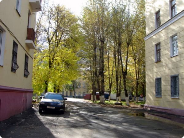 Черный автомобиль припаркован на асфальтированной дороге между двумя желтыми жилыми домами, окружёнными высокими деревьями с желто-зелёной листвой в солнечный день.