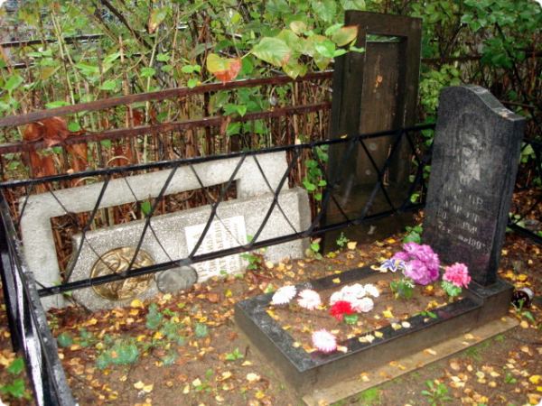 Надгробие с портретом и цветами на фоне других памятников и ржавой решётки среди осенних листьев.