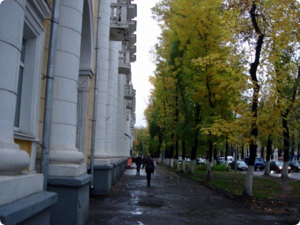 Прогулка по осенней аллее у белого здания с колоннами: люди идут по мокрому тротуару, деревья в жёлто-зелёной листве, на заднем плане — припаркованные машины.