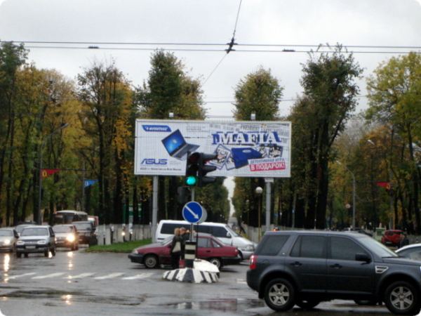 Большой рекламный щит с надписью «МАРИЯ» и логотипом ASUS на фоне осенних деревьев и мокрой дороги с автомобилями и светофором.