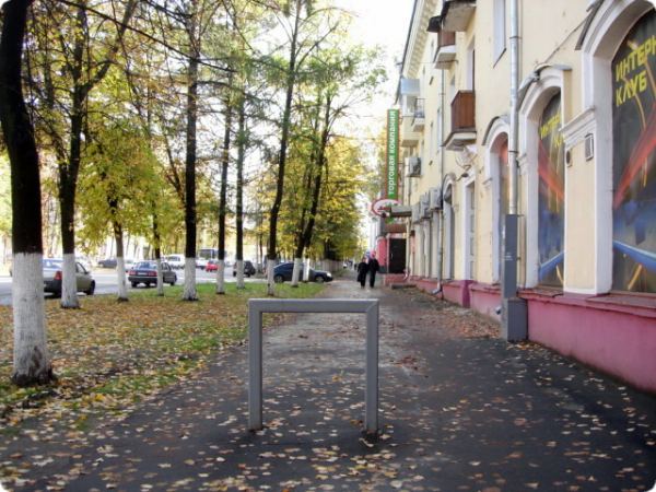 Осенняя улица с деревьями, покрытыми жёлтыми листьями, и металлической рамой на тротуаре; справа — здание с вывеской «Интер Кафе», слева — парковка с автомобилями.