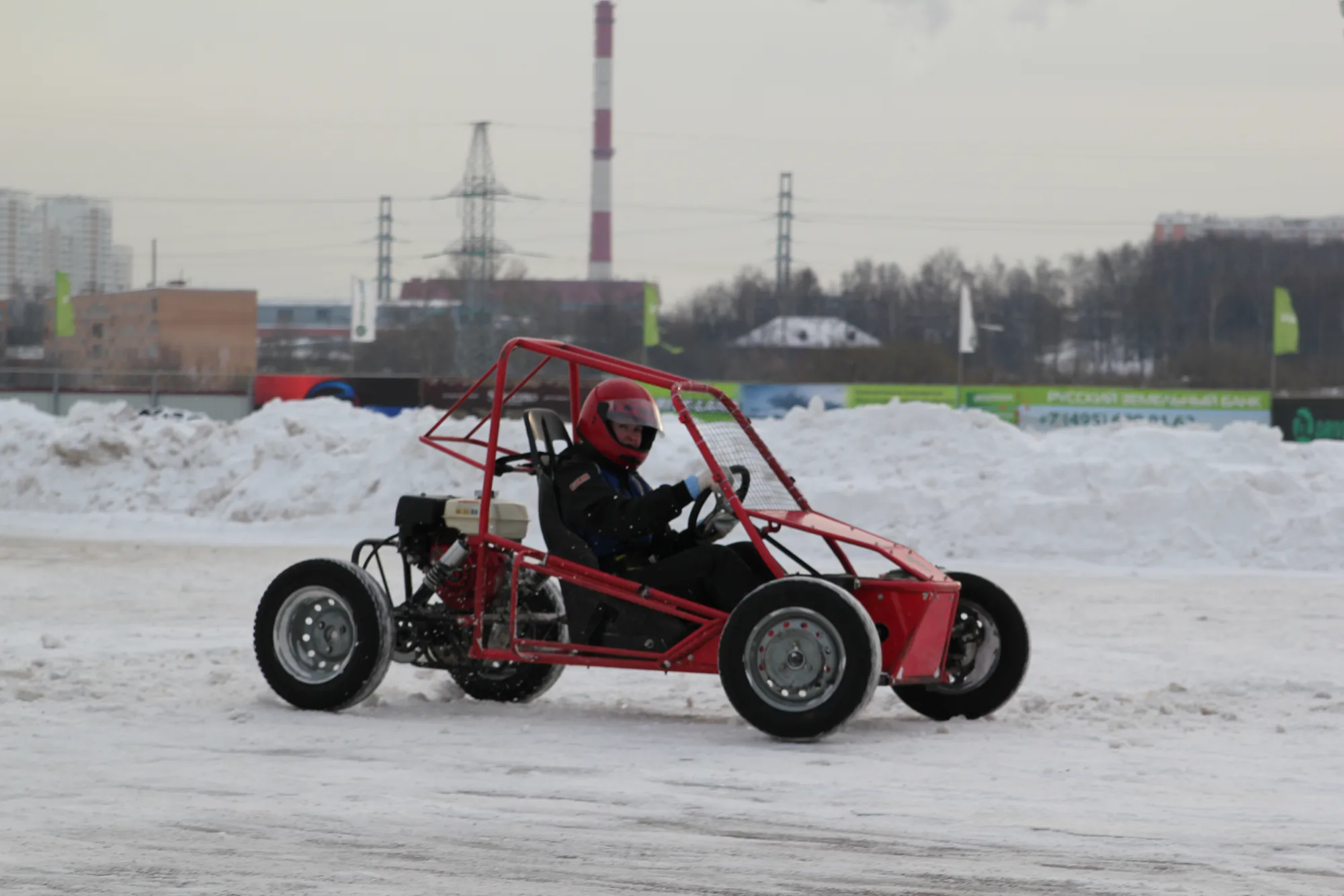 Человек в красном шлеме управляет красным гоночным багги по заснеженной трассе на фоне промышленных зданий и труб.