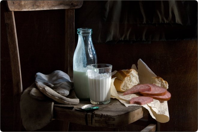 Стеклянная бутылка и стакан молока, ломтики хлеба и колбасы на деревянном стуле, рядом сложенная ткань и ложка.