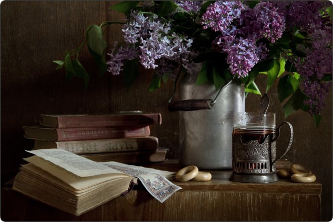 Натюрморт: раскрытая книга, стопка старых книг, букет сирени в металлическом кувшине, стакан чая в подстаканнике и сушки на деревянном столе.