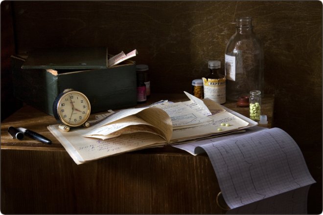 На деревянном столе лежат старинные медицинские документы, часы, перьевая ручка, стеклянные флаконы с лекарствами и пилюлями, создавая атмосферу прошлого.