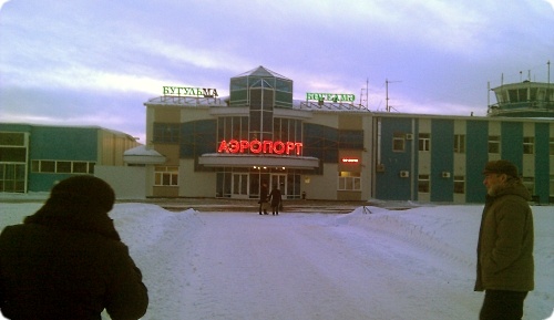 Зимний вид здания аэропорта Бугульма с красной неоновой вывеской «АЭРОПОРТ» и зелёными надписями «БУГУЛЬМА» на крыше; на переднем плане — люди в зимней одежде на снегу.