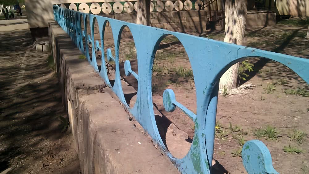 Синяя металлическая ограда с круглыми вырезами на бетонном парапете в парке, на заднем плане видны деревья и декоративная стена с круглыми элементами.