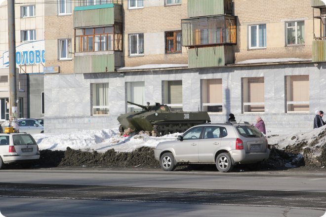 Зелёная боевая машина пехоты с номером 318 стоит на снежном валу у здания с вывеской «ОЛОВО», рядом припаркованы легковые автомобили и проходят пешеходы.