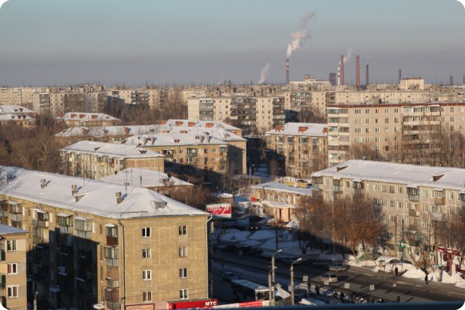 Зимний городской пейзаж: многоэтажные жилые дома с заснеженными крышами, улицы с автомобилями и пешеходами, на горизонте — промышленные трубы с дымом.