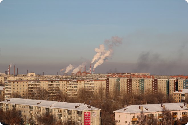 Промышленные трубы выбрасывают дым над жилыми многоэтажными домами в зимнем городе, покрытом снегом.