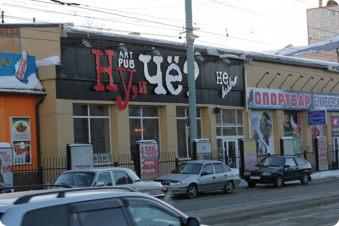 Фасад здания с вывеской «Ну, и чё?» (арт-паб) и «Спортбар Букмекерский», припаркованные машины, снежные крыши, уличные фонари.