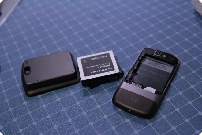 Разобранный смартфон HTC Desire: задняя крышка, аккумулятор 3000 мАч и корпус с пустым отсеком для батареи на синей клетчатой поверхности.