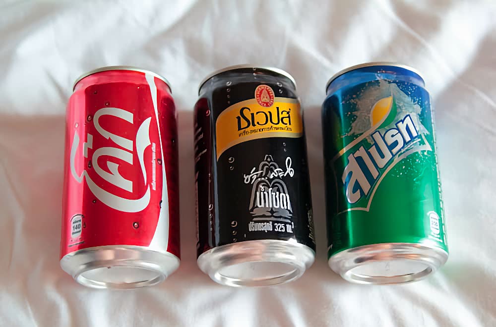 Три банки газированных напитков с тайскими надписями: красная Coca-Cola, чёрная энергетик и зелёная Sprite, лежат на белой ткани.