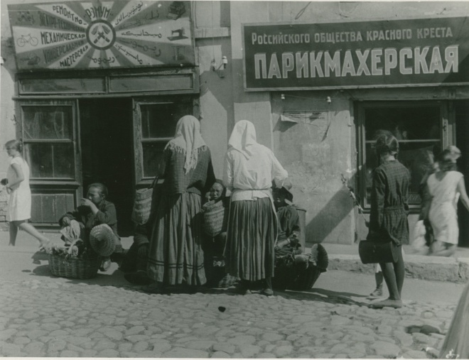 Чёрно-белое фото: женщины в платках и дети у лавок с вывесками «Парикмахерская» и «Механическая мастерская», люди сидят на тротуаре, одна девочка идёт мимо.