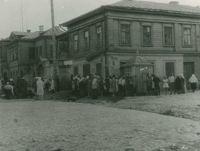 Чёрно-белая фотография: длинная очередь людей у деревянного здания с крыльцом и вывеской, на фоне других домов и пыльной улицы; видны женщины в платках, дети, мужчина с тележкой.