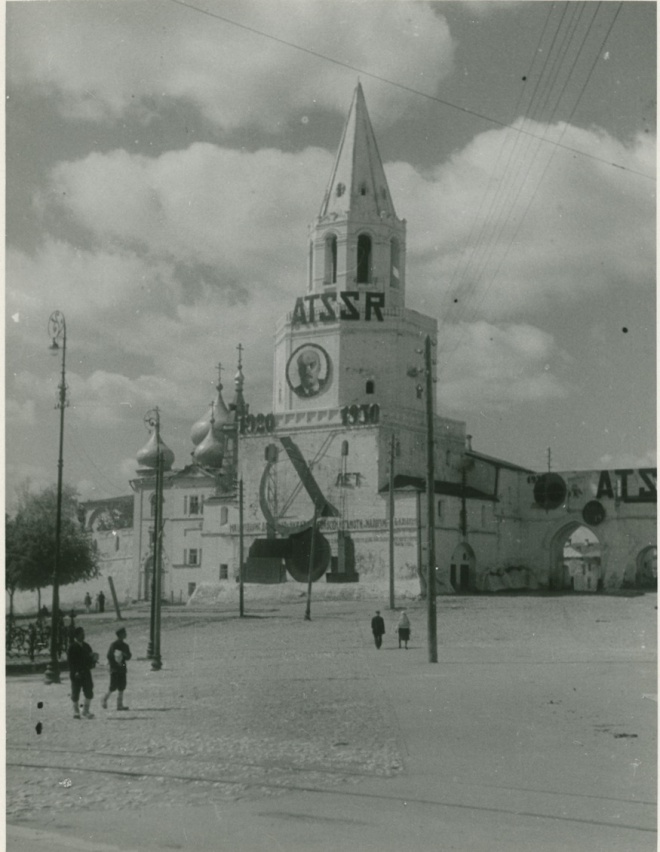 Чёрно-белое фото: башня с портретом Ленина, надписью «АССР» и датой 1930, рядом — купола церкви, люди на площади, облачное небо.