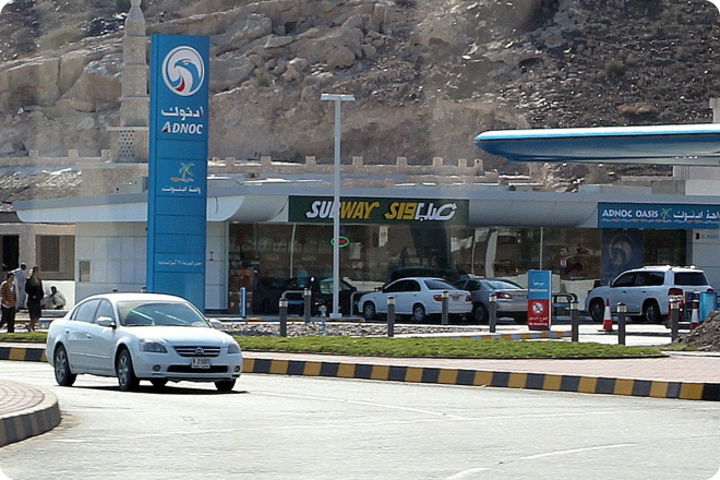 Заправка ADNOC Oasis с рестораном Subway, белая машина на переднем плане, скалистый склон на заднем плане.