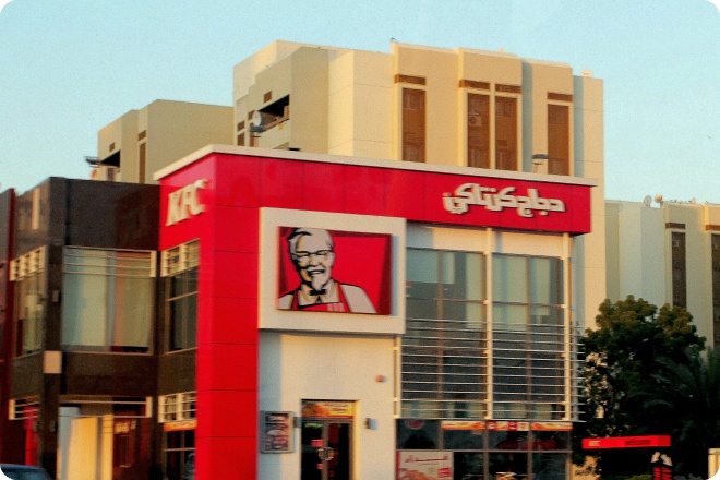 Фасад ресторана KFC с красной вывеской, на которой изображён полковник Сандерс и арабская надпись «KFC