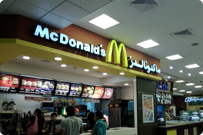Вход в ресторан McDonald's с вывеской на английском и арабском языках, меню на экранах и посетители у кассы