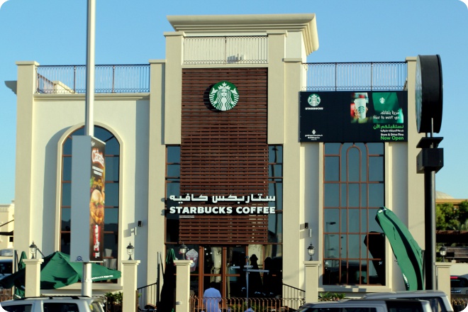 Фасад здания Starbucks с вывеской на арабском и английском языках, зелёным логотипом и рекламным баннером справа; видны вход, окна, зонты и автомобили на переднем плане.