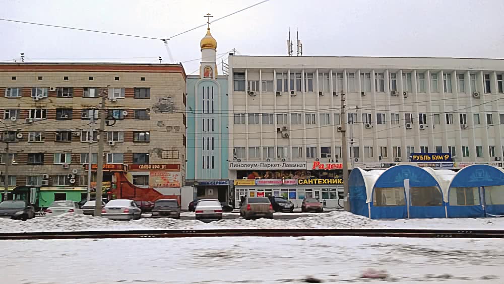 Зимний городской пейзаж: многоэтажные здания с магазинами, церковь с золотым куполом, припаркованные машины и снежная дорога на переднем плане.