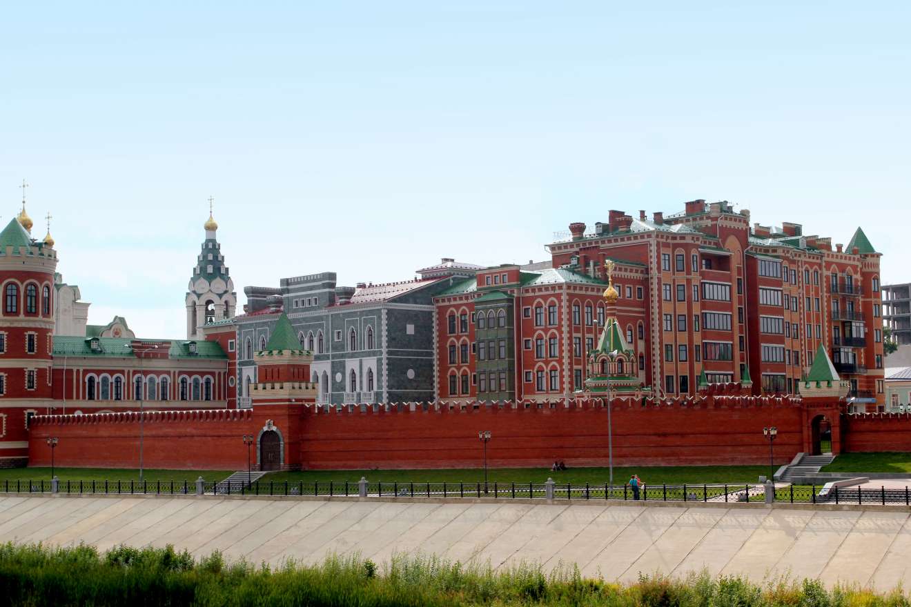 Красная кирпичная стена с башнями и зубцами, за ней — многоэтажные здания в стиле исторического центра с золотыми куполами и зелёными крышами на фоне ясного неба.