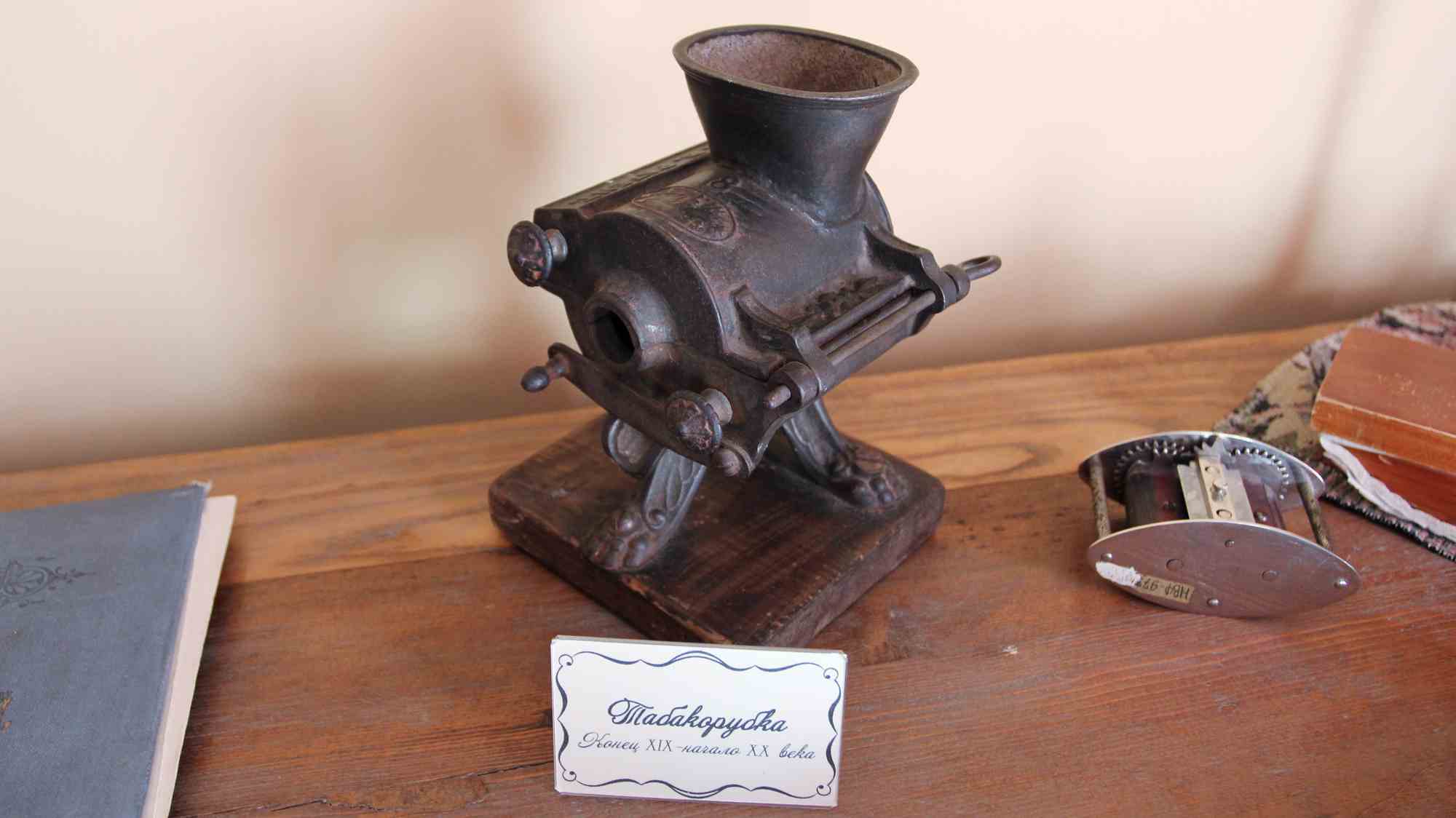 Старинная металлическая табакорубка на деревянной подставке с этикеткой «Табакорубка. Конец XIX – начало XX века», рядом — разобранный механизм и книги на столе.