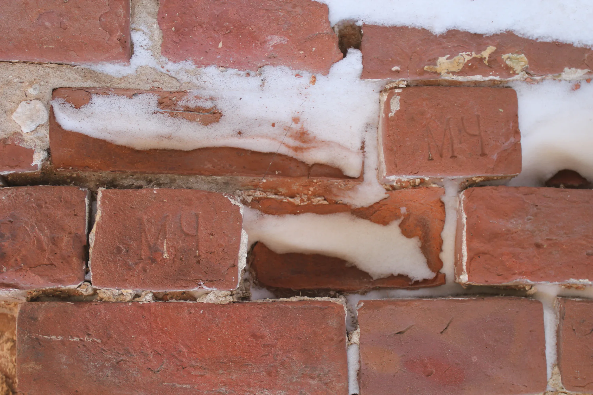 Крупный план старой кирпичной кладки с остатками снега в швах; на двух кирпичах видны выбитые буквы «МЧ».