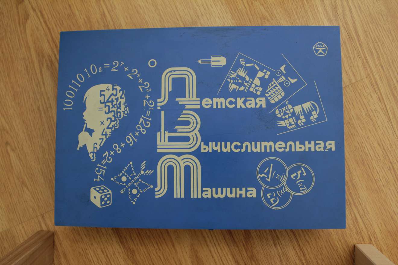 Синяя коробка с надписью «Детская Вычислительная Машина», логотипом СССР, математическими формулами, силуэтом головы и игральными костями на деревянном фоне.