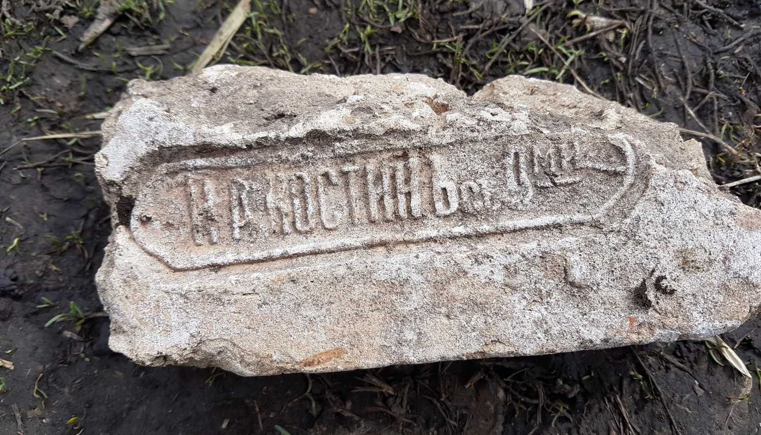 Старый кирпич с оттиском «А. А. Костинъ» в овальной рамке, лежащий на влажной земле среди травы и веток.