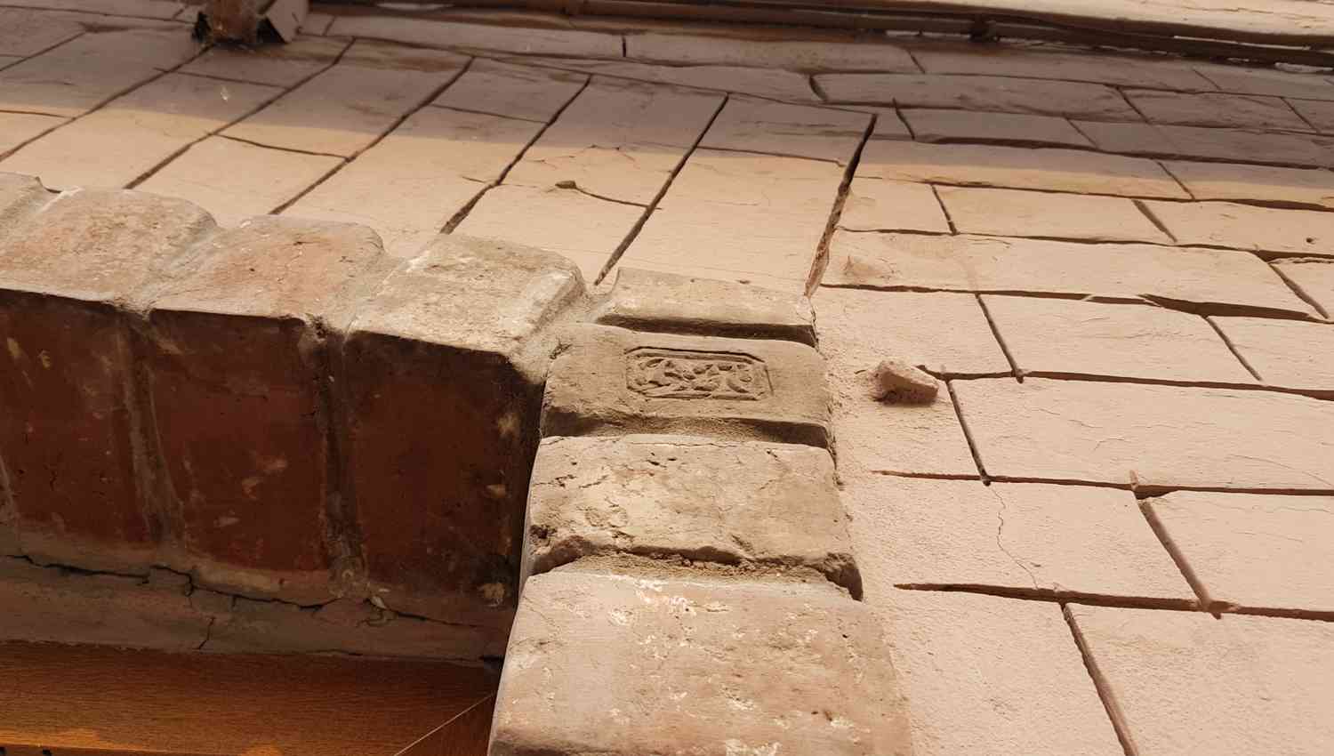 Старинная кирпичная кладка с декоративным рельефным узором на одном из кирпичей, уложенная на фоне светлых каменных плит.