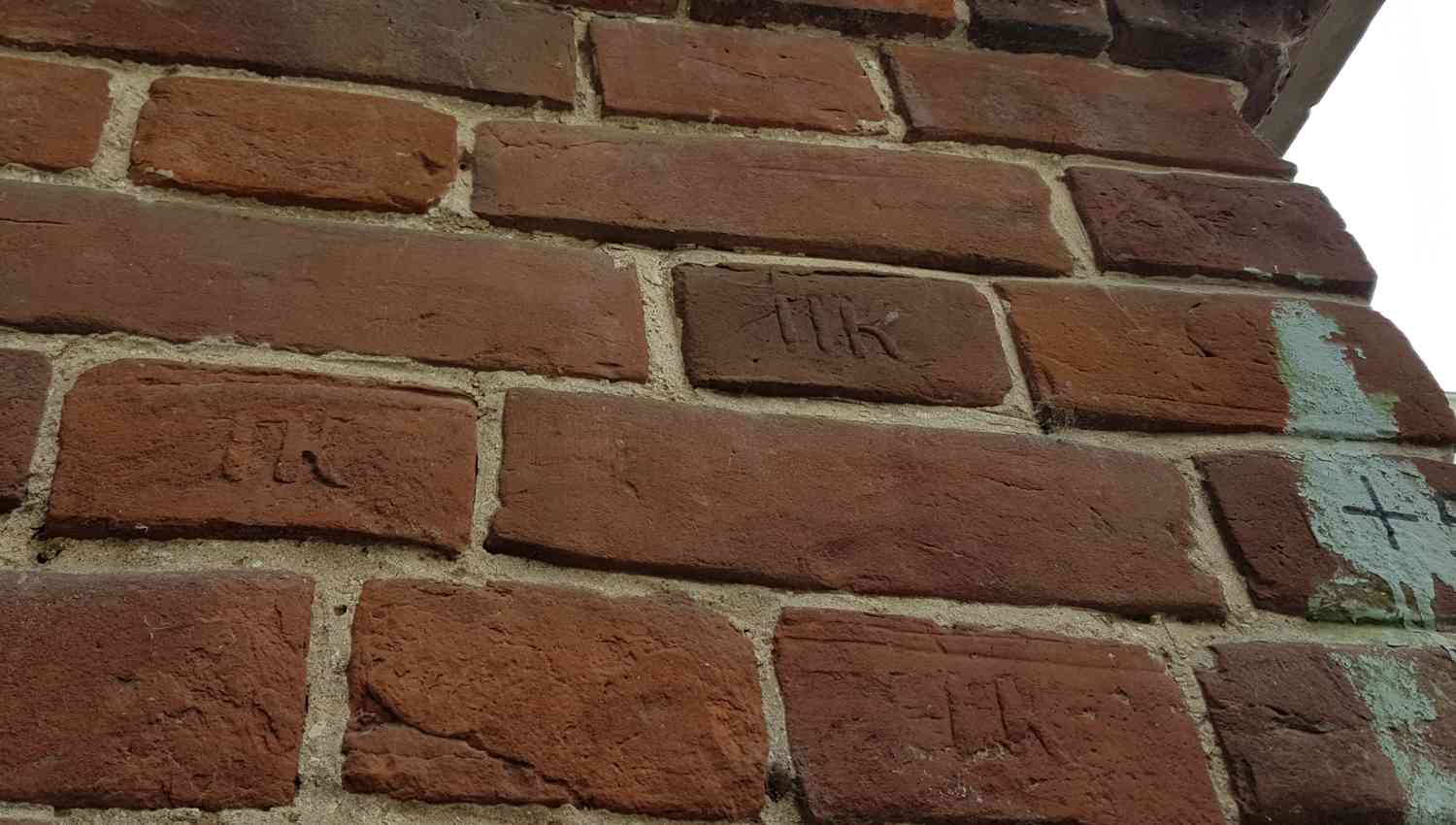Крупный план красной кирпичной кладки с вытесанными на некоторых кирпичах буквами «ПК» и участками старой зеленой краски с крестом.