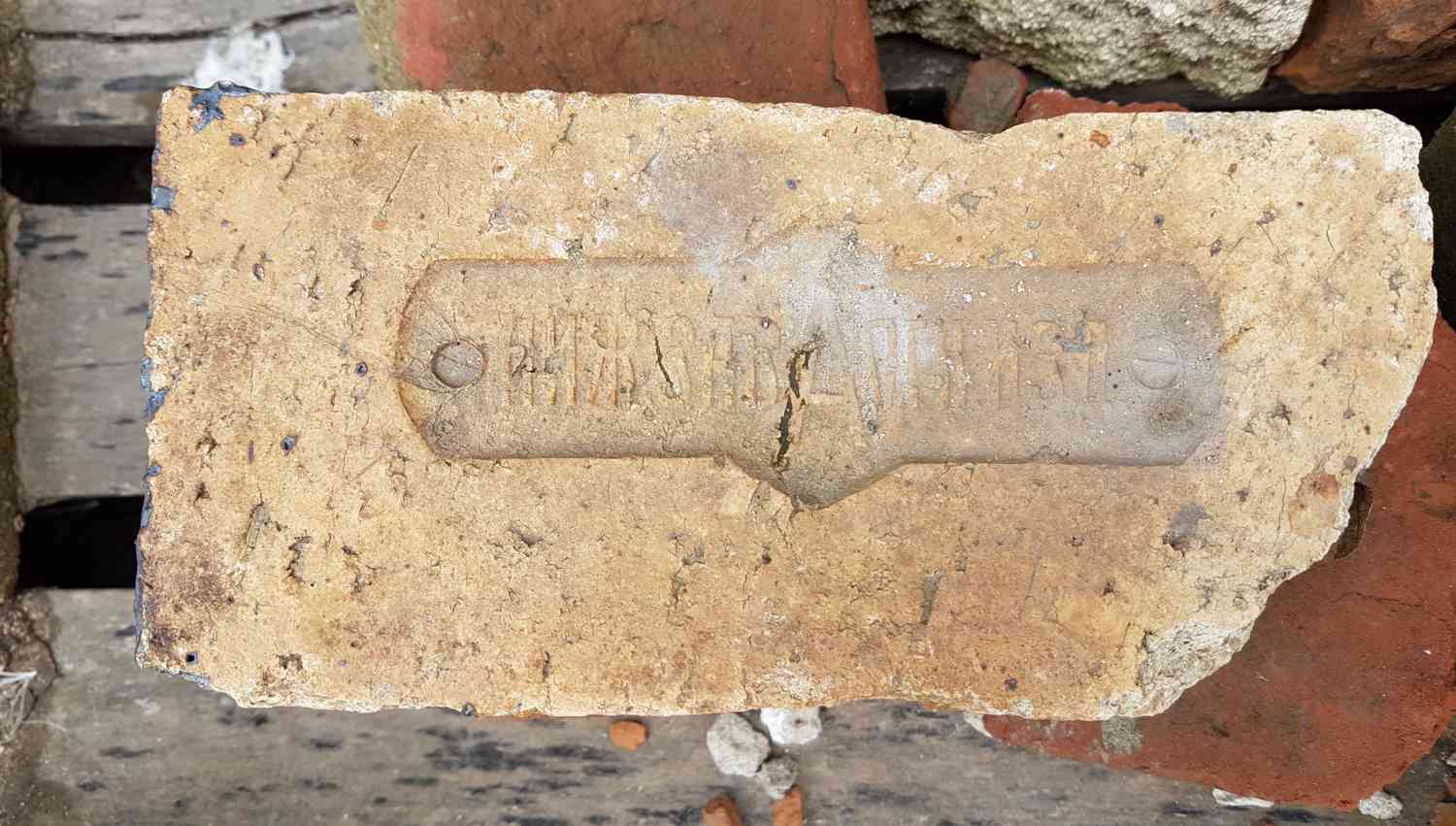 Старая кирпичная плитка с выгравированным логотипом завода «Красный Октябрь» и номером партии, лежащая среди строительного мусора.