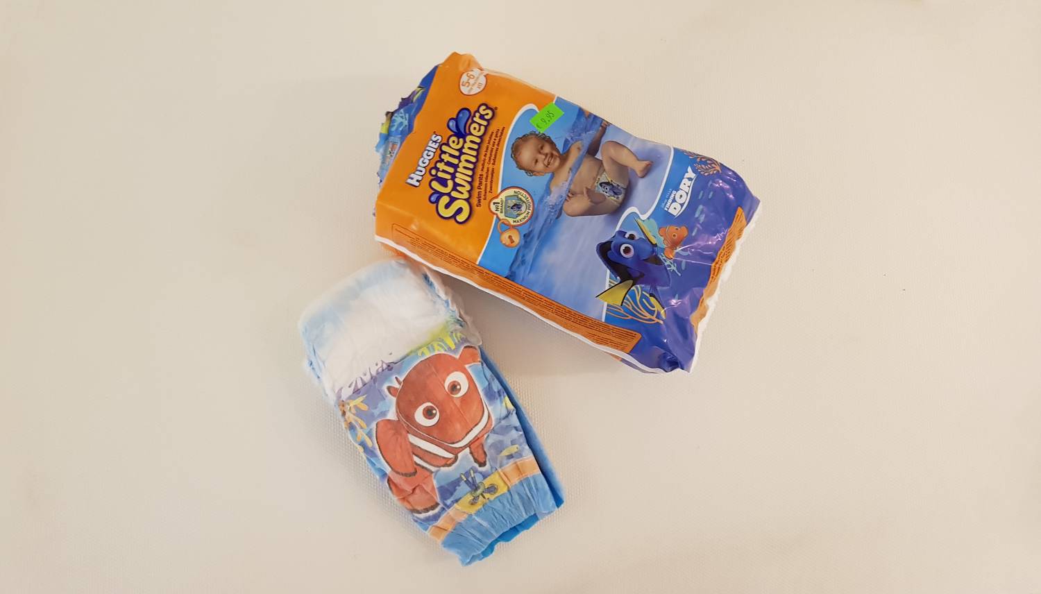 Пакет подгузников Huggies Little Swimmers с персонажами «В поисках Дори» и один вынутый подгузник с изображением рыбки-клоуна на светлом фоне.