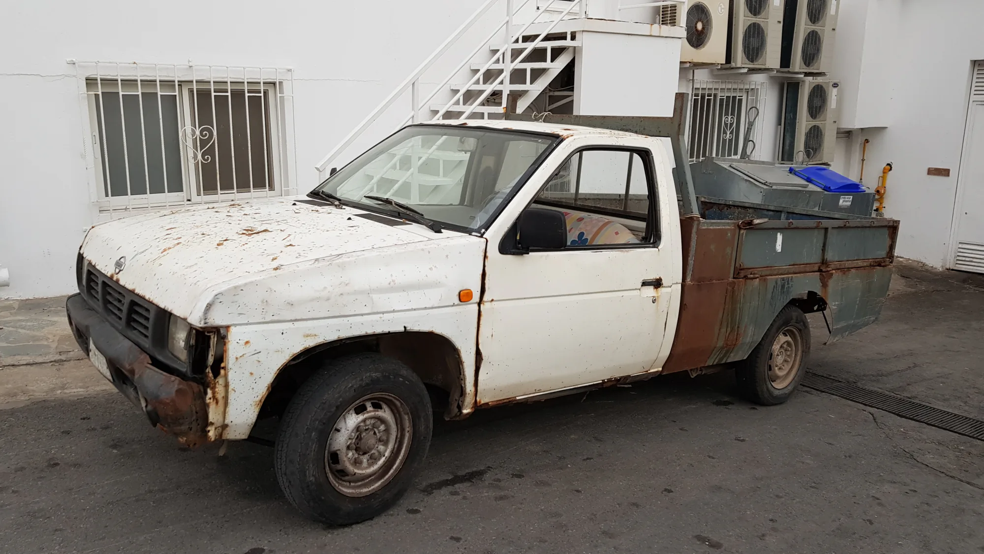 Ржавый белый пикап Nissan с зелёным кузовом и синим контейнером на заднем плане у белого здания с кондиционерами и лестницей.