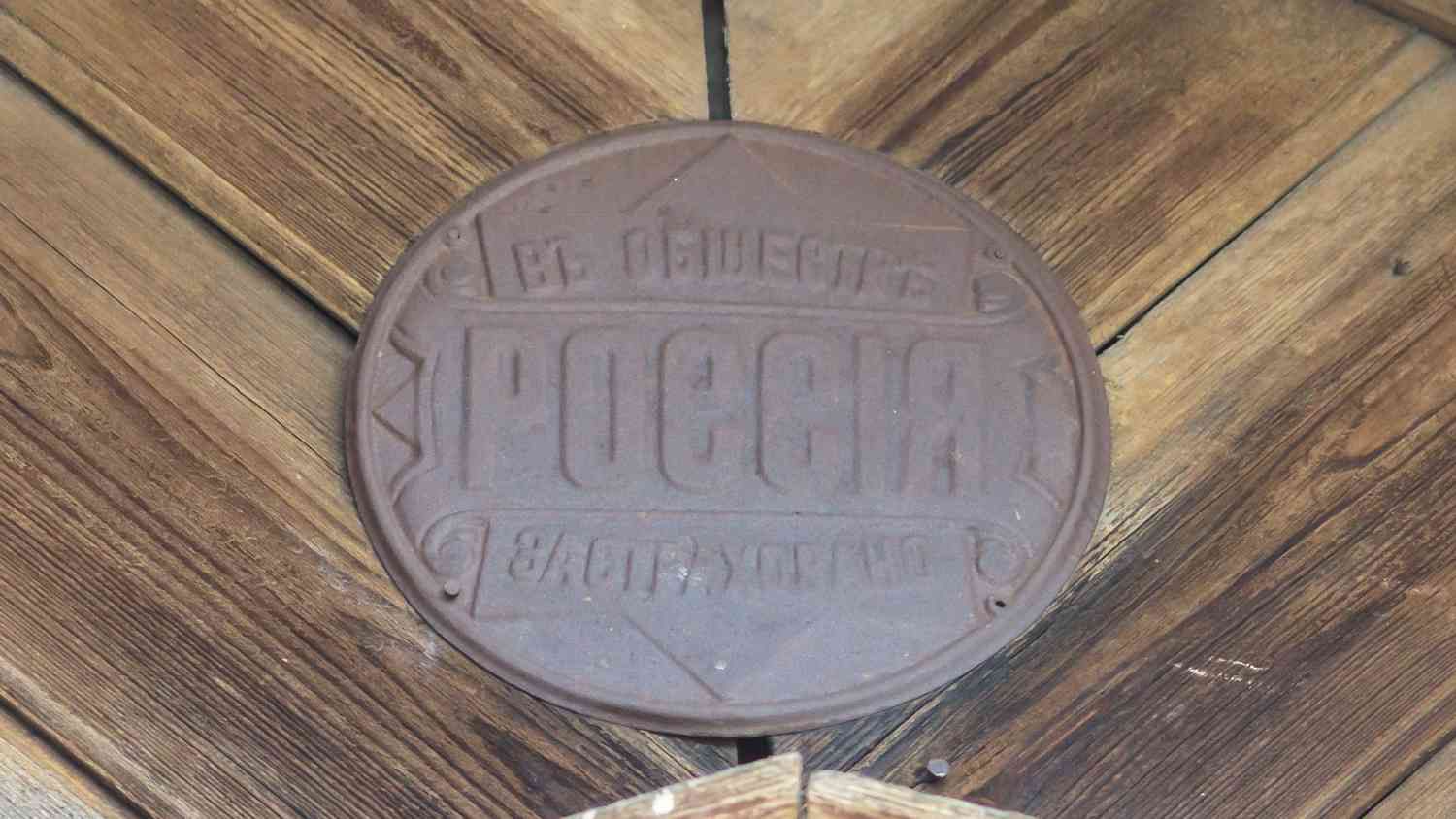 Круглая металлическая табличка с надписью «РОССИЯ» и словами «В ОБЩЕСТВЕ» и «В СТРАХЕ БОЖИЕМ» на деревянном полу.