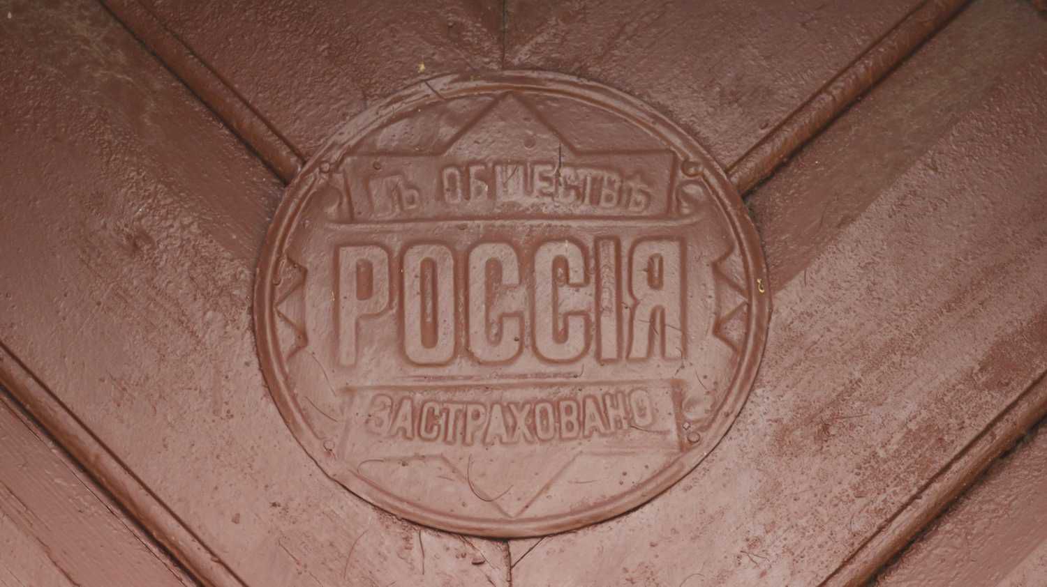 Круглая металлическая эмблема на деревянной поверхности с надписью «ОБЩЕСТВО РОССІЯ ЗАСТРАХОВАНО» в стиле начала XX века.