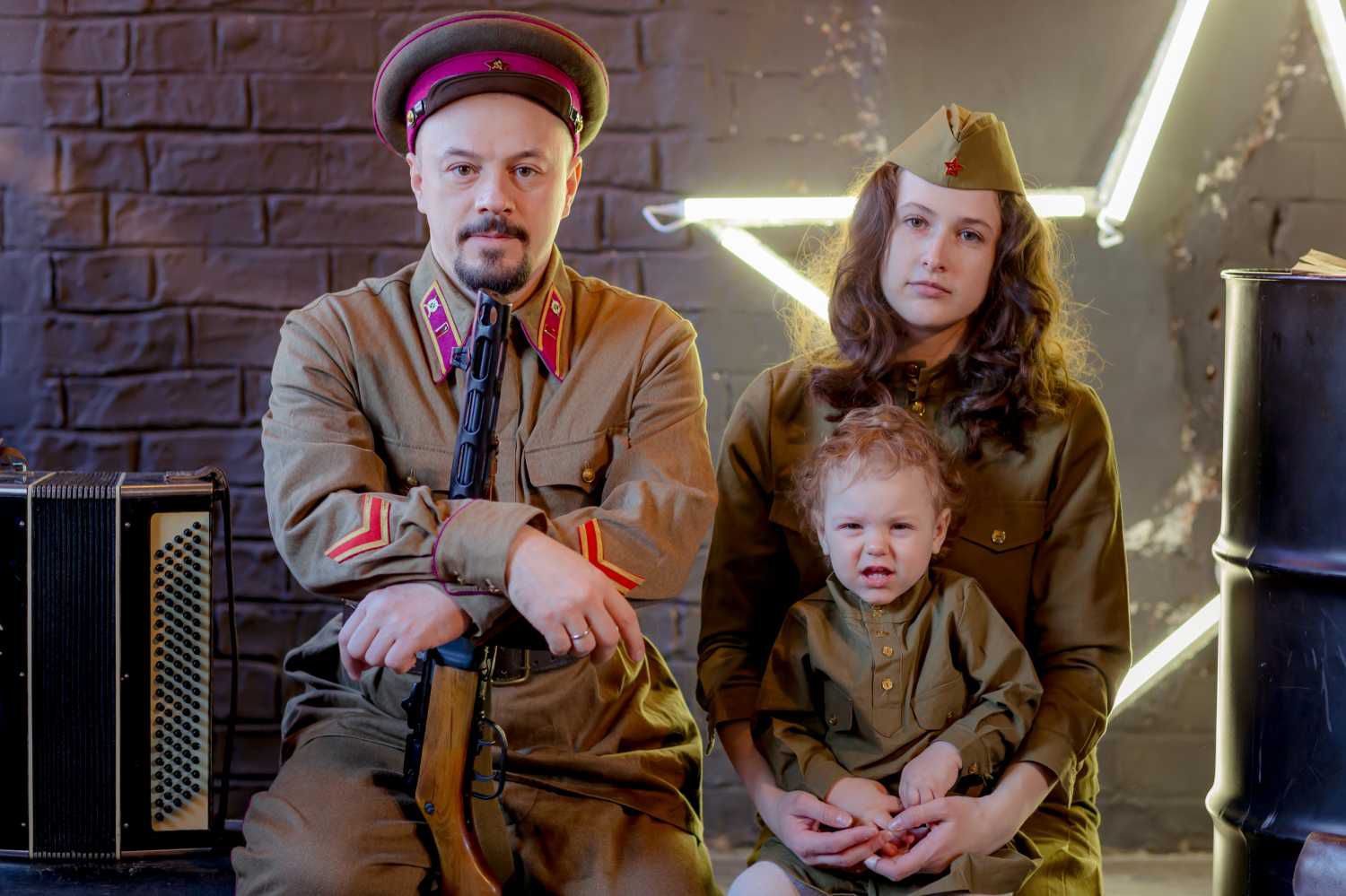 Семья в советских военных костюмах: мужчина с винтовкой и аккордеоном, женщина с ребёнком на фоне кирпичной стены и неоновой звезды.