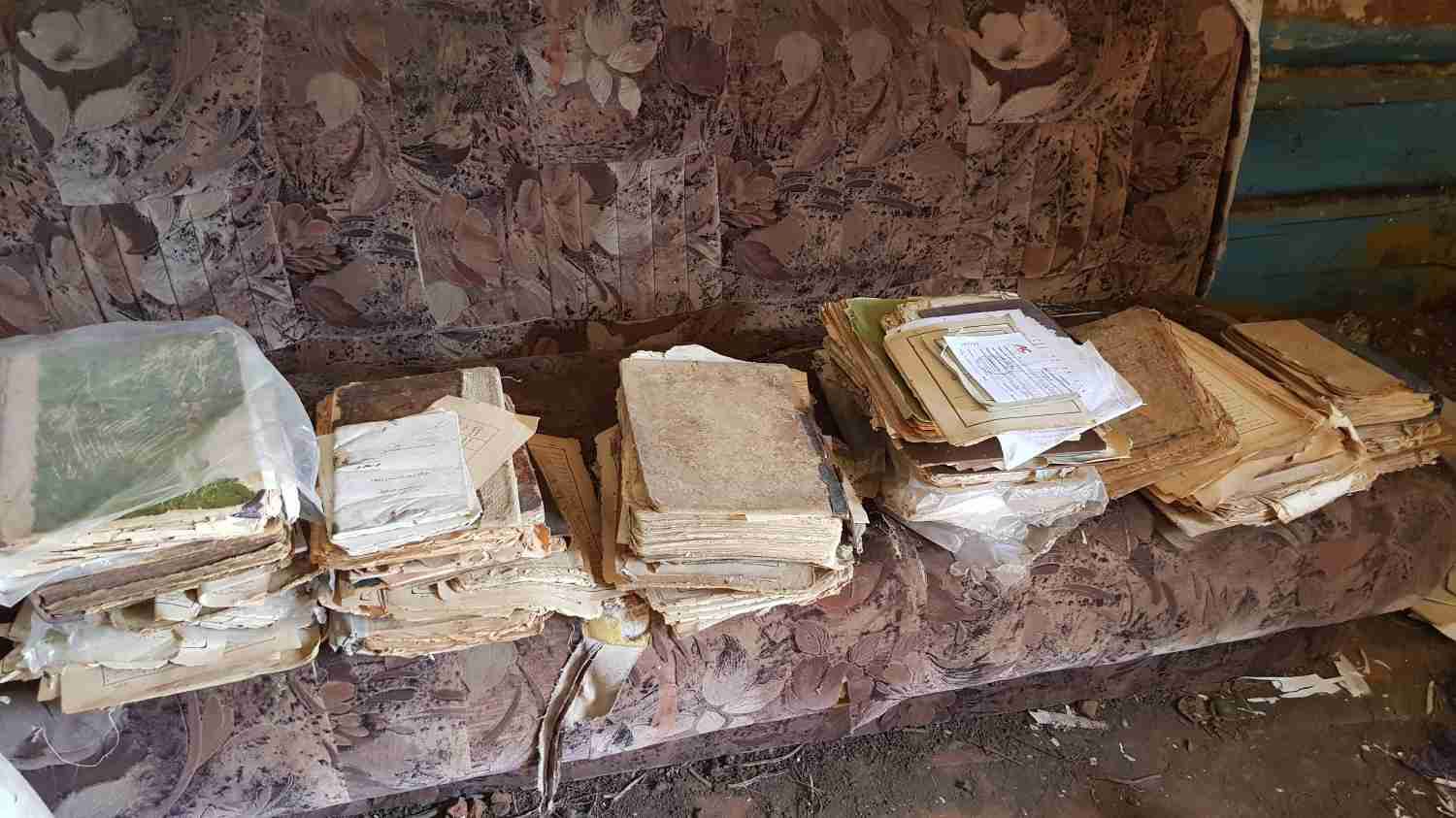 Сгоревшие архивные документы и папки, сложенные в стопки на старом диване с цветочным узором, часть из них обёрнута в прозрачный пластик.
