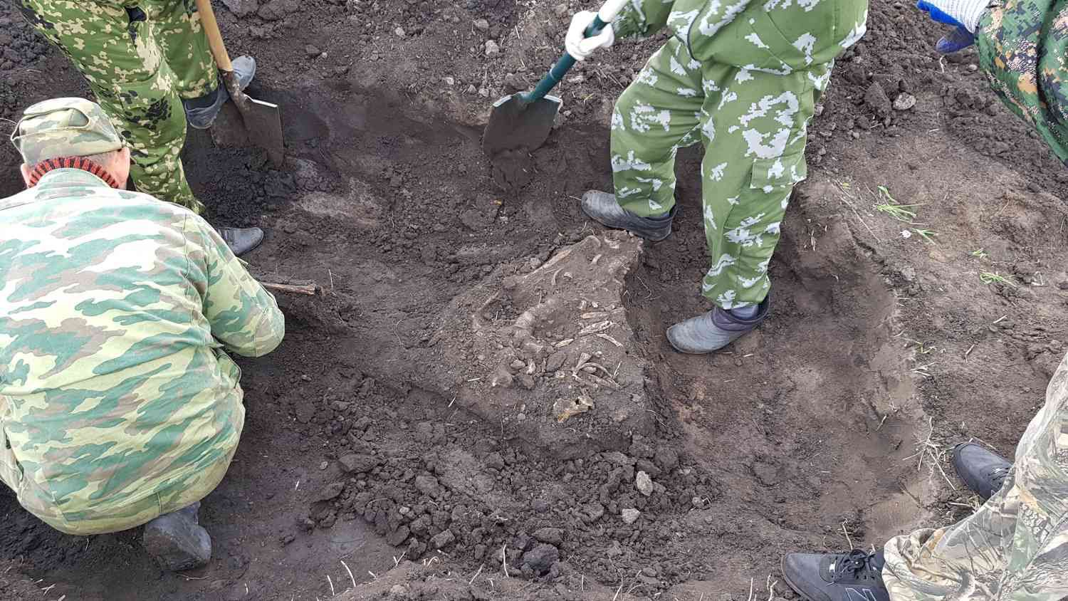 Люди в камуфляже раскапывают лопатами и кистями останки человека в земле, проводя археологические или следственные работы на открытом грунте.