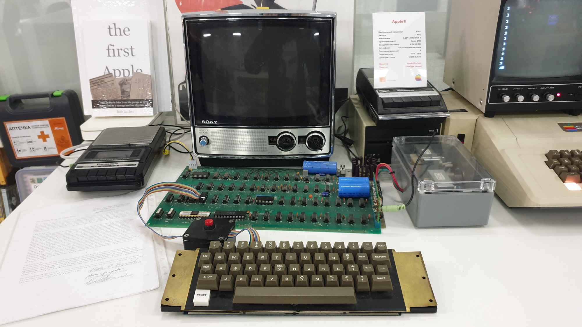 Экспонат Apple II: материнская плата, клавиатура, кассетный магнитофон, монитор Sony и информационная карточка с характеристиками компьютера на столе.