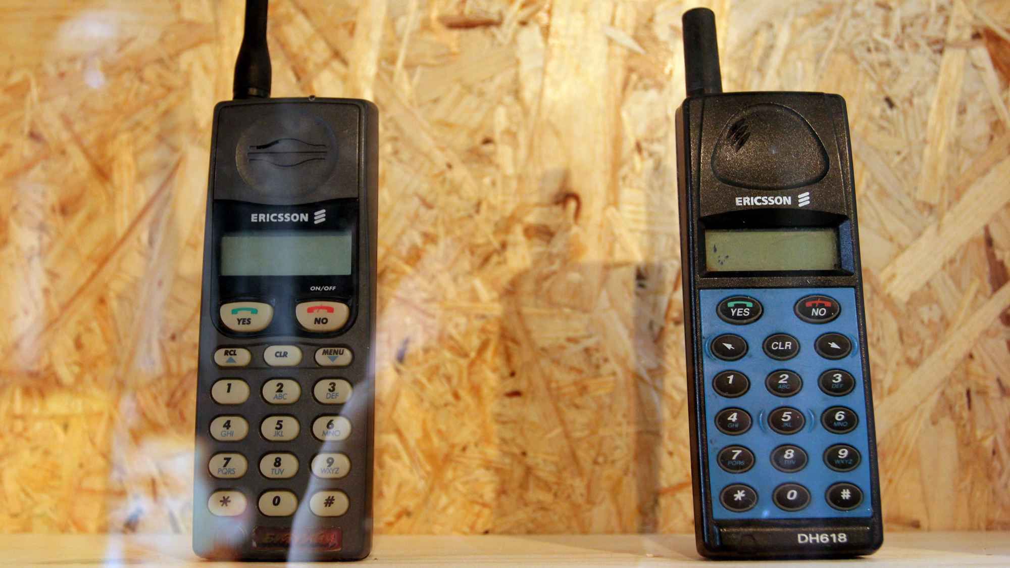 Два чёрных мобильных телефона Ericsson с антеннами на деревянной панели: слева — модель с серыми кнопками, справа — DH618 с синей клавиатурой и надписью YES/NO.