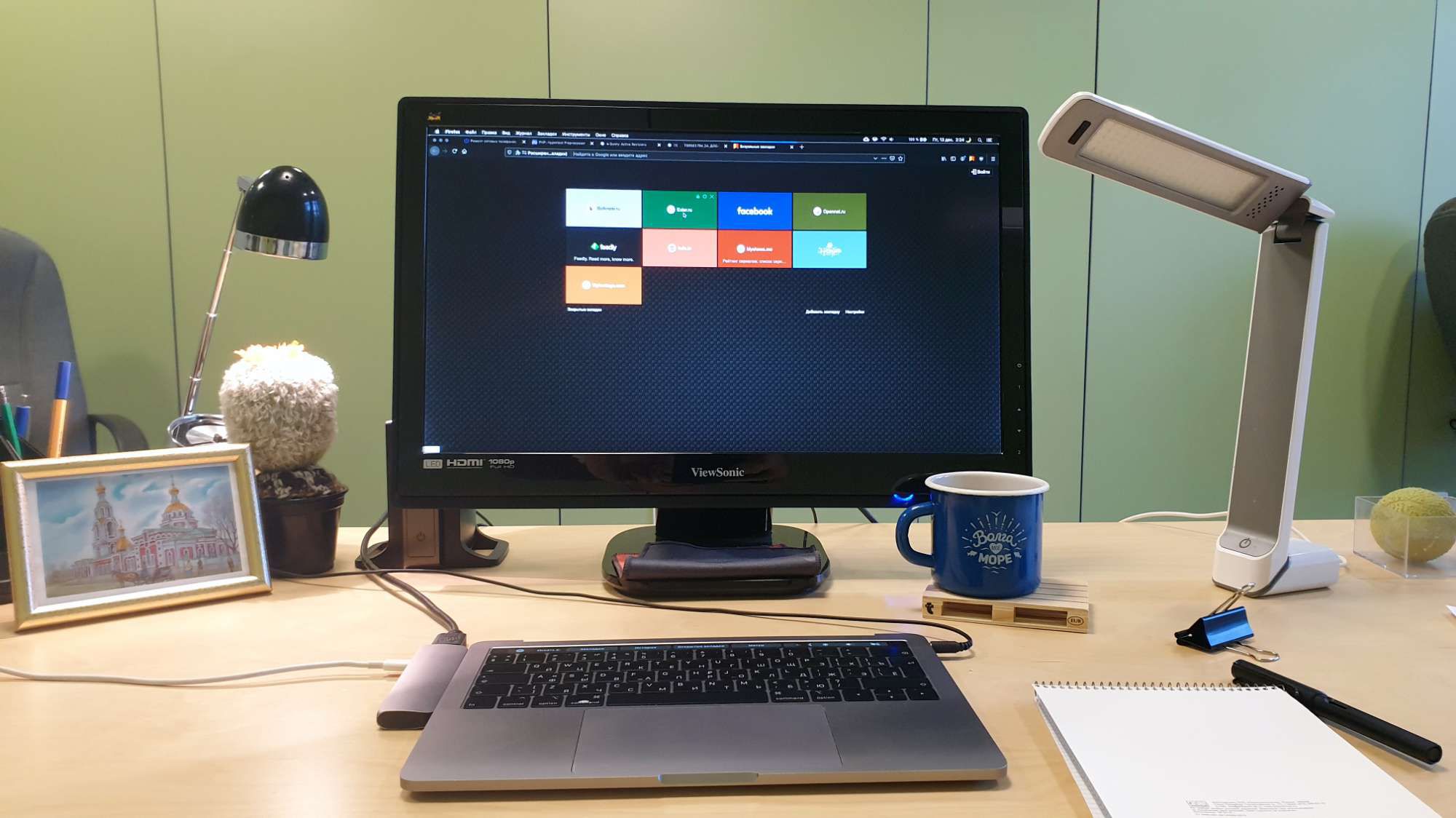 Рабочий стол с ноутбуком, монитором ViewSonic, двумя лампами, синей кружкой, блокнотом, рамкой с картиной и кактусом на светлом столе.