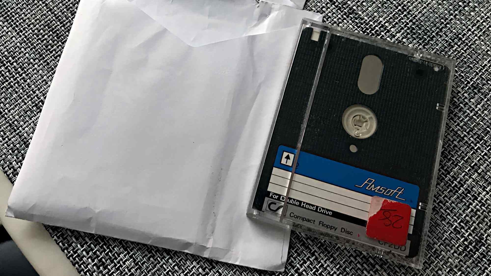 Компакт-диск Amsoft в прозрачном футляре с надписями «For Double Head Drive», «Compact Floppy Disc» и красной наклейкой «28» лежит на белой бумаге и серой плетёной поверхности.