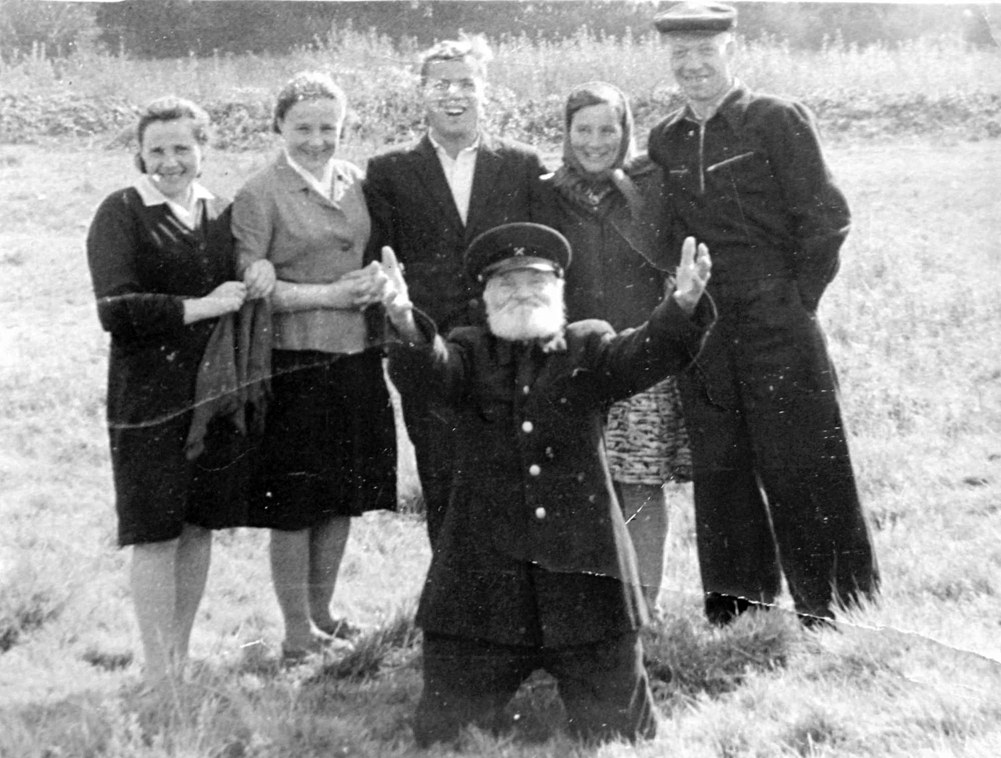 Чёрно-белое фото: пожилой мужчина с белой бородой в форме и фуражке стоит на коленях, подняв руки, за ним — пятеро улыбающихся молодых людей на фоне травы.