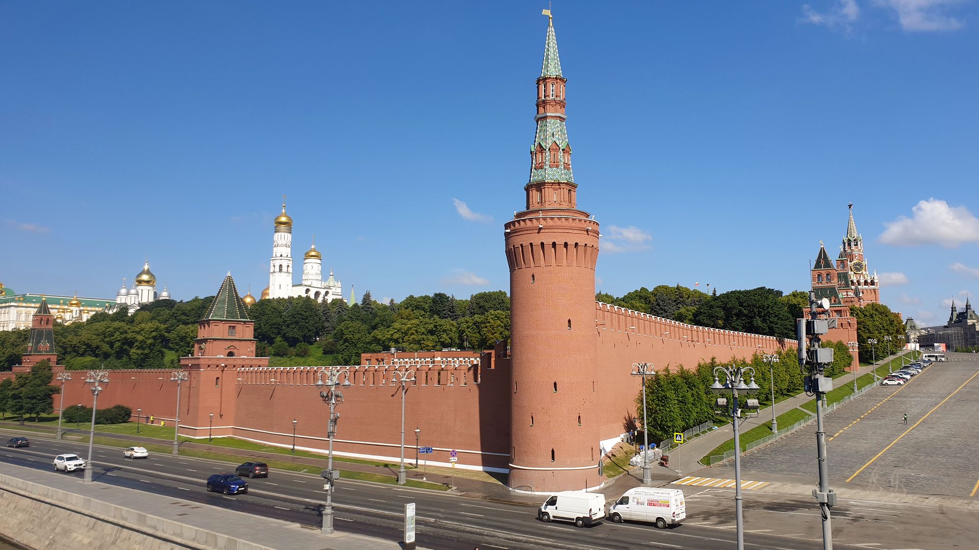 Вид на Московский Кремль: красная кирпичная стена, Боровицкая башня с зелёной шатровой крышей, золотые купола соборов и Спасская башня на фоне ясного голубого неба.